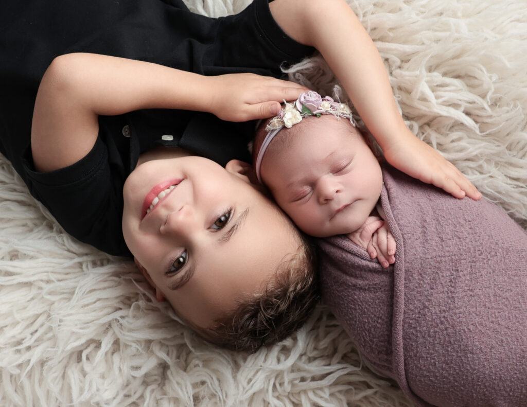 A heartwarming photo of a big brother gently laying beside his newborn sister, captured in a cozy setting at MJ Memories Photography studio in Rochester, NY.