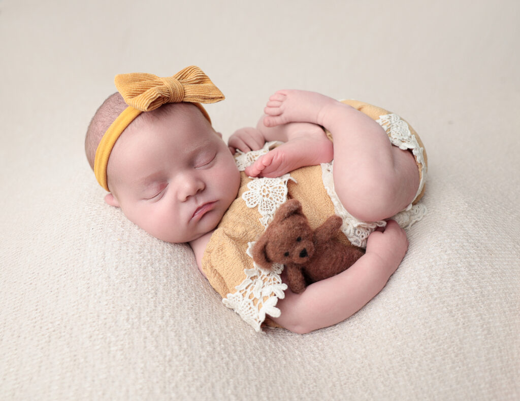 Newborn girl dressed in a gold outfit, gently holding a teddy bear, captured in a cozy photography studio.