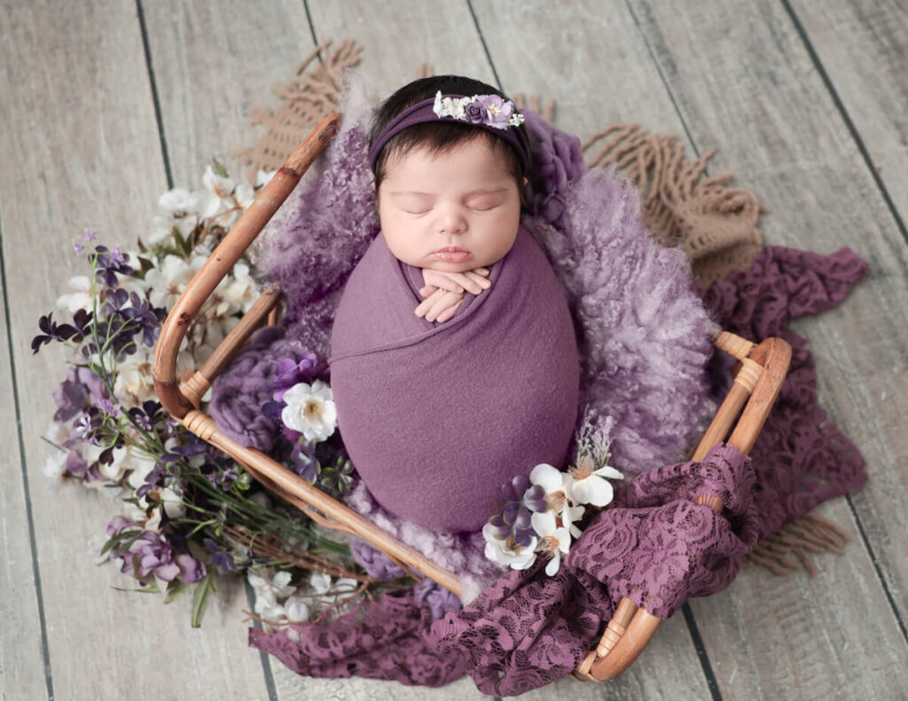 A beautiful newborn baby girl surrounded by purple florals, captured at MJ Memories Photography studio in Rochester, NY.