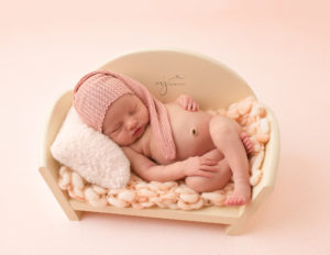 Baby girl in a ivory bed laying on a pillow and wearing a pink sleepy cap at her newborn photography session 