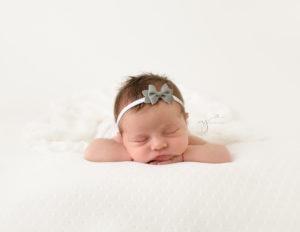 Baby girl in head on hands newborn photography pose on a white backdrop wearing a bow headband