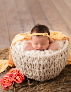 Newborn in a cream basket in head on hands pose wearing a yellow headband and draped with a yellow wrap