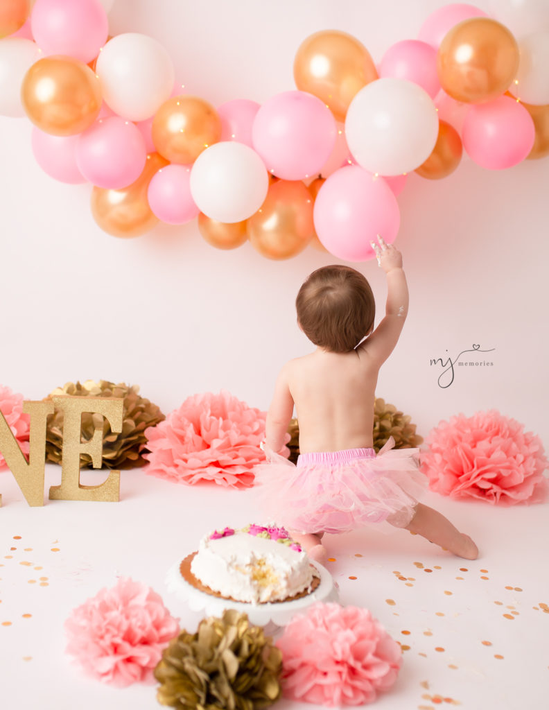 Backside of a one year old girl pointing at pink gold white balloons at her cake smash photography session