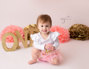 Baby girl smiling on a white backdrop with paper flowers and gold letters ONE