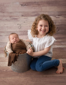 Big sister sitting next to her newborn brother who is wrapped in a brown wrap in metal bucket